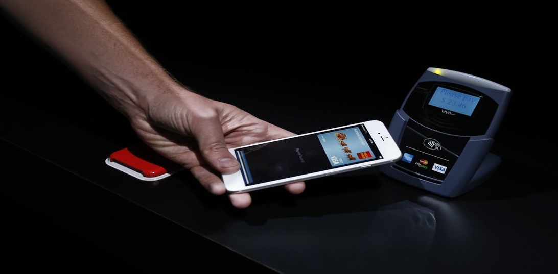 En hand håller en smartphone nära en kontaktlös betalterminal och visar Apple Pay i användning med Touch ID-autentisering.